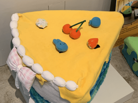 <b>Felt Cake Model</b><br>The model originates from the Sheridan Crafts and Design building.In this soft cake model, the bright yellow fabric top resembles icing, while the white pom-poms create a decorative border. The playful use of colorful felt toppings mimics real cake decorations, enhancing the model's whimsical appeal. The combination of textures, from the soft felt to the smooth surfaces, invites touch and interaction.