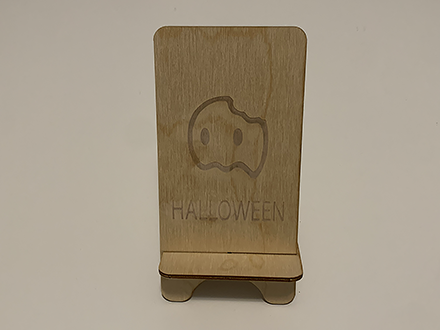 <b>Front View of the Phone Stand</b><br>
          This is the front view of the phone stand I made in the Sheridan laser cutting room. It has a Halloween theme with a cute ghost engraving and the word 'HALLOWEEN' underneath. The design is simple and fits well on the wood surface.