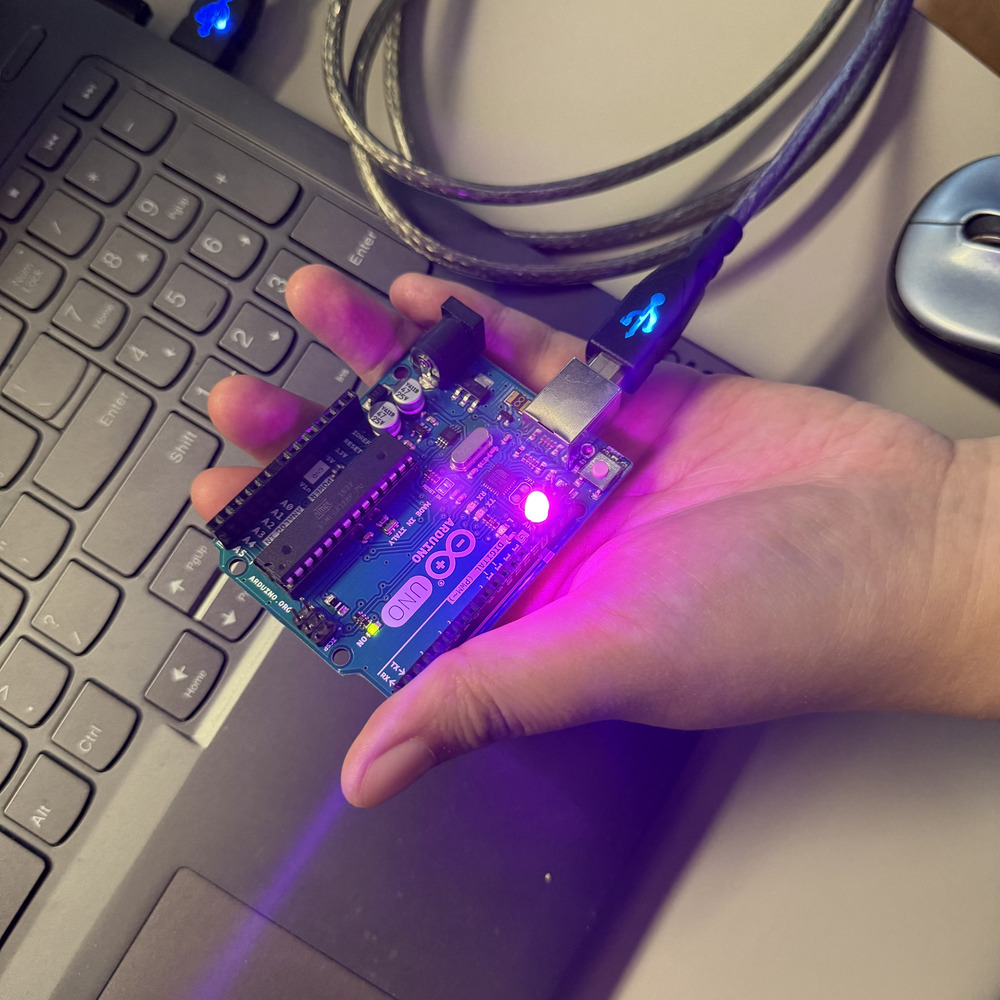 <b>Powering the Arduino UNO</b><br>I am holding an Arduino UNO board that is connected to my laptop. The board is powered through a USB cable, and a bright purple onboard LED is glowing. This tells me that the Arduino is working correctly and ready for programming.